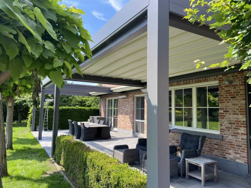 Overdekt terras met tuinmeubilair aan een bakstenen huis, omgeven door gras en bomen.