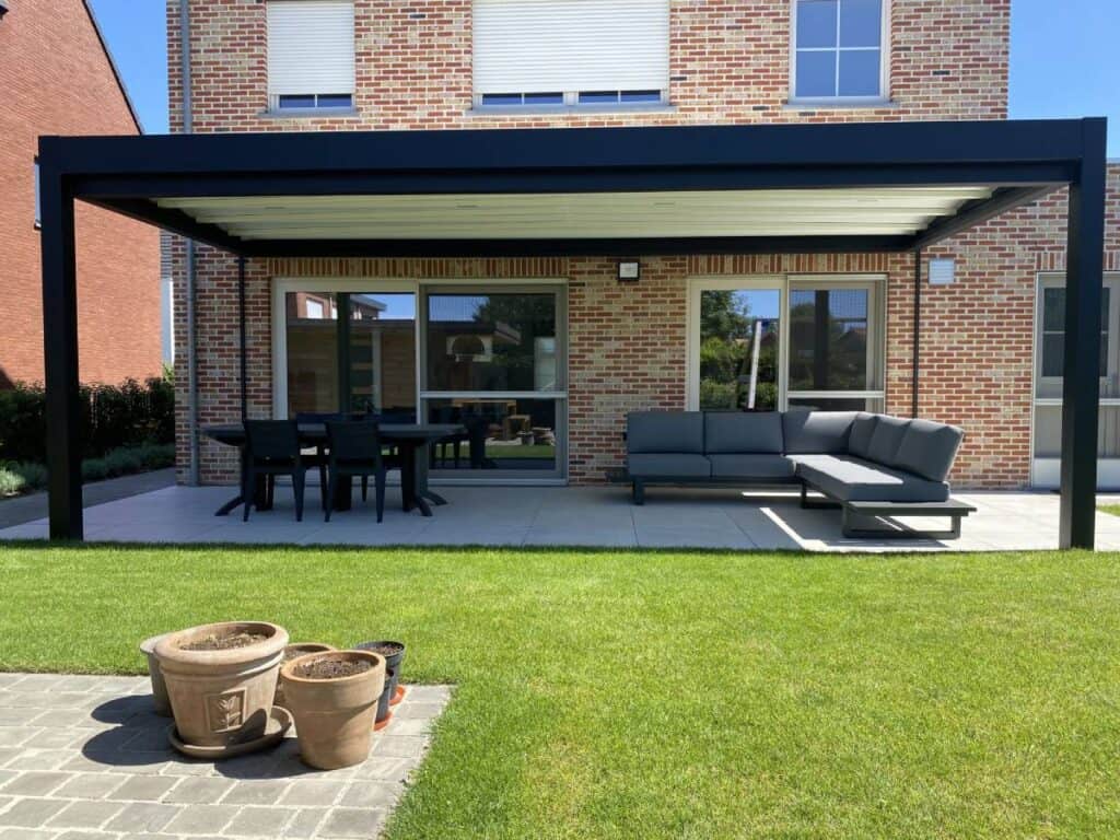 Terras met overkapping, hoekbank, eettafel met stoelen en bloempotten op betegeld stuk gras.