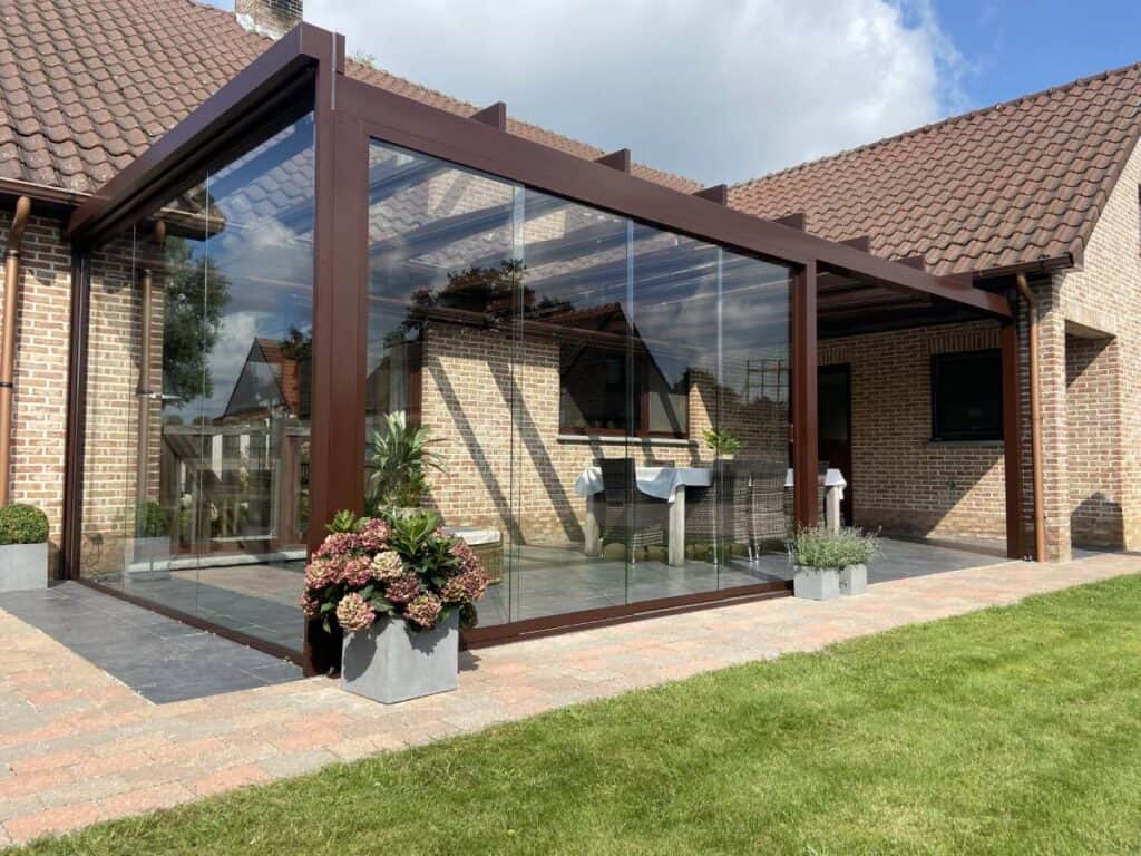 Huis met glazen serre, betegeld terras, planten in potten en grasveld eromheen.