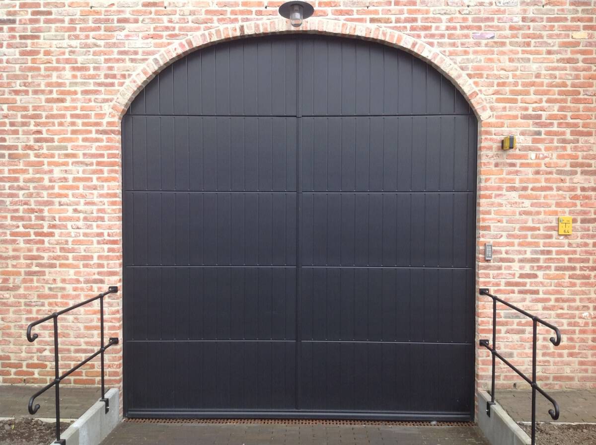 Zwarte dubbele garagepoort met verticale panelen in rode bakstenen boogmuur, metalen leuningen.