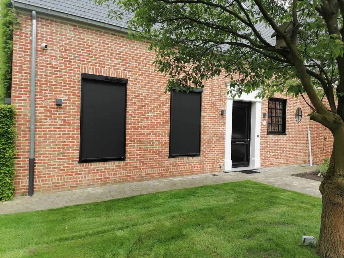Gevel van een bakstenen huis met zwarte screens, een zwarte deur met wit kozijn en een boom in de voortuin.