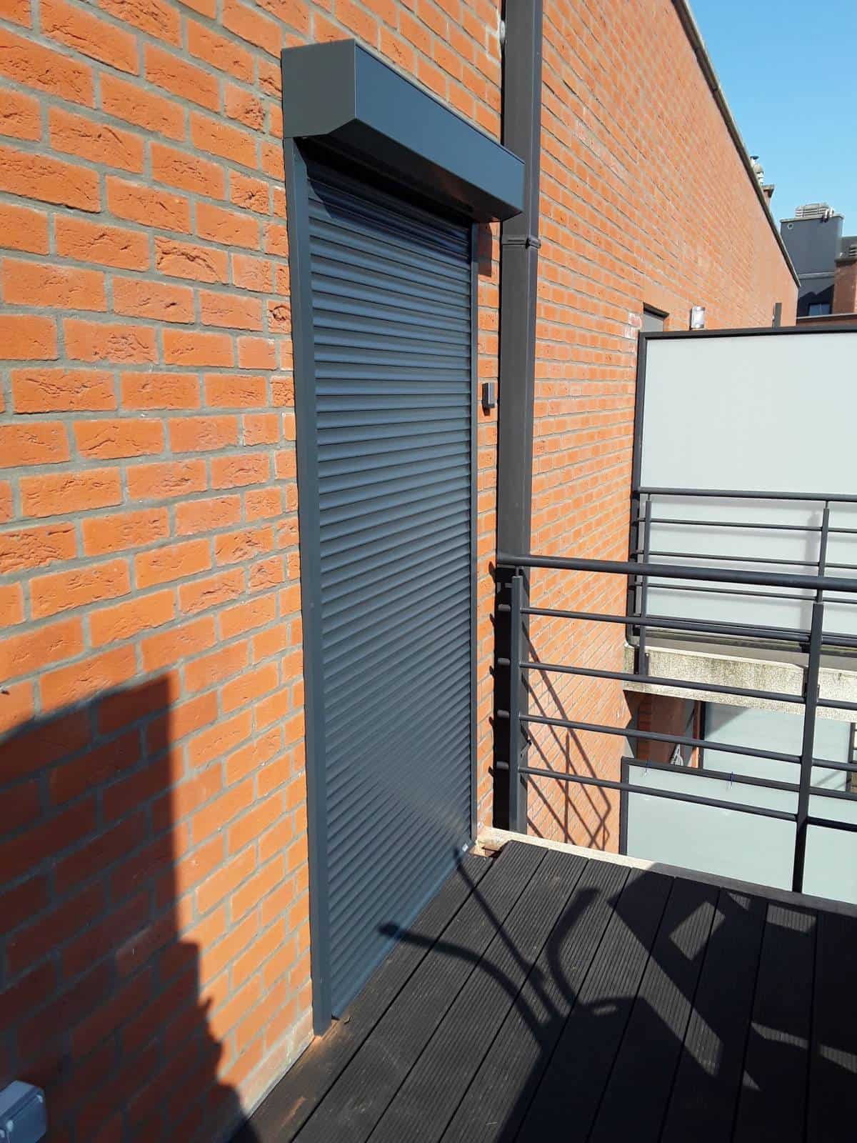 Balkon met houten vloer, metalen balustrade met matglazen panelen en gesloten grijze rolluikdeur.