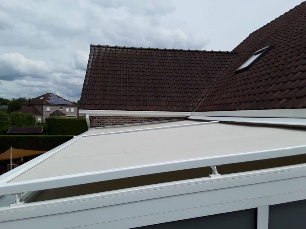 Dak met donkere dakpannen en wit Verandazonwering op overkapping, bewolkte lucht en huizen op achtergrond.