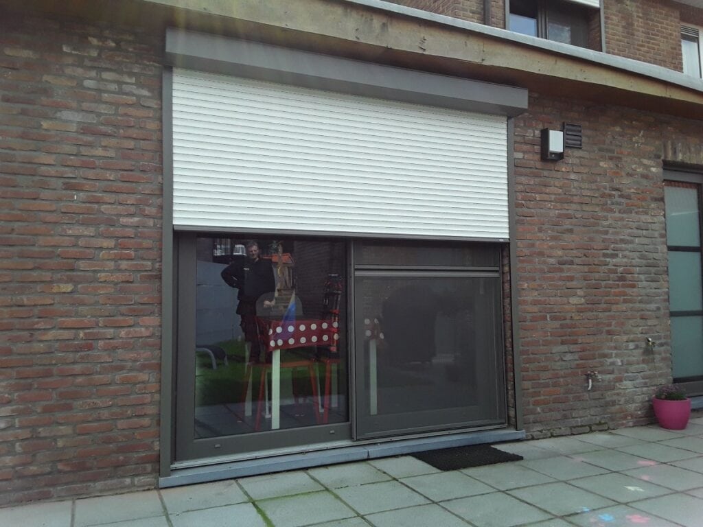 Glazen schuifdeur met deels gesloten wit rolluik, matje voor deur en roze bloempot tegen bakstenen muur.