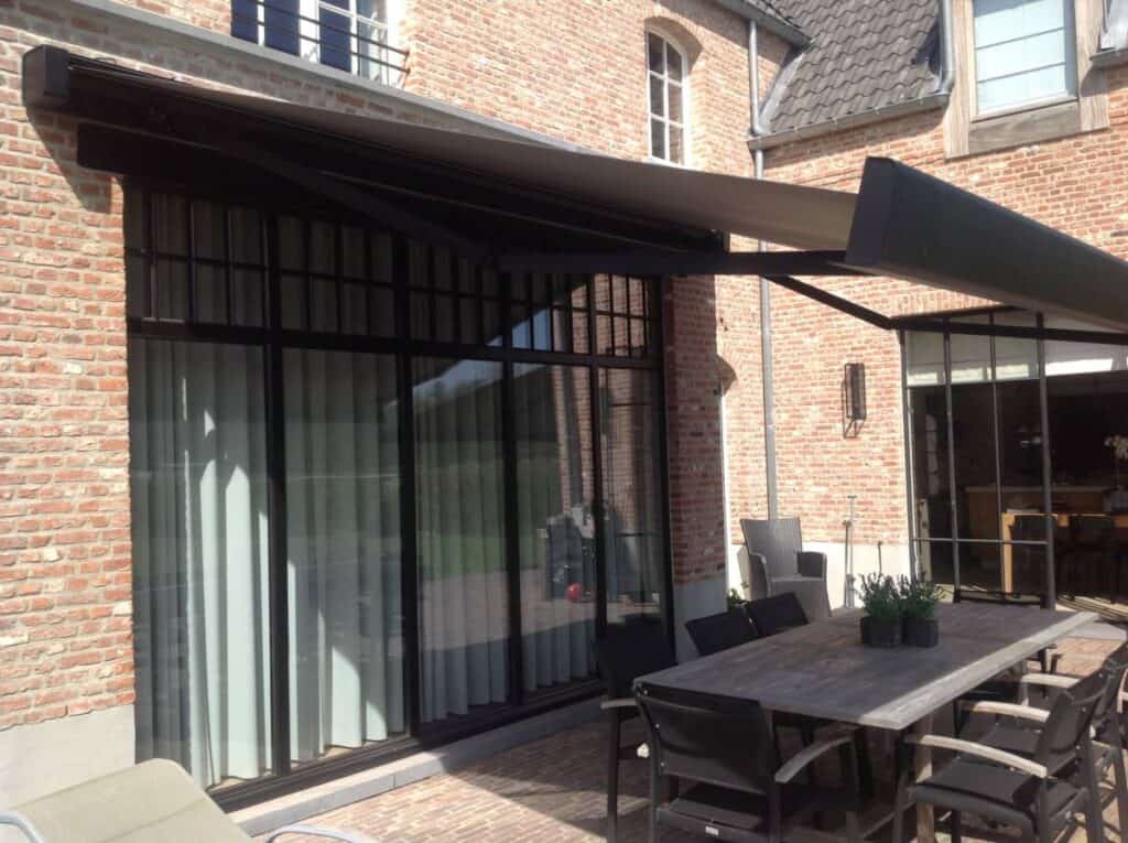 Houten eettafel met zes zwarte stoelen onder zonnetent naast bakstenen gebouw met grote ramen.