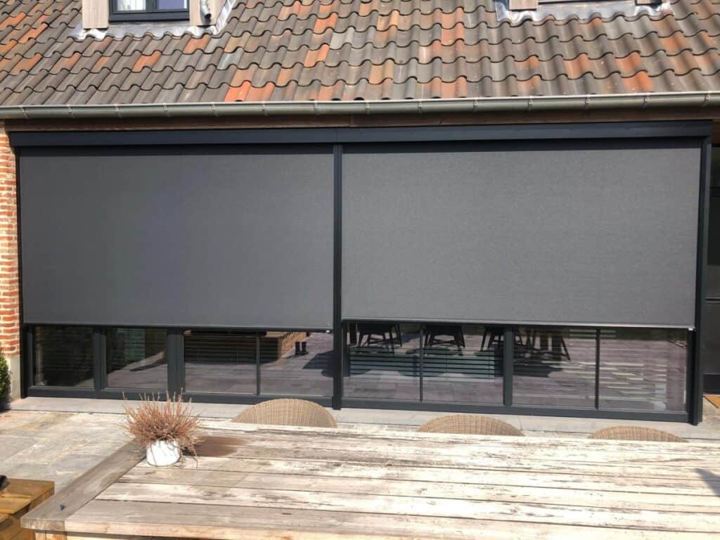 Huis met dakpannen, grote ramen met grijze screens, houten tuinset en pot met gedroogde planten.
