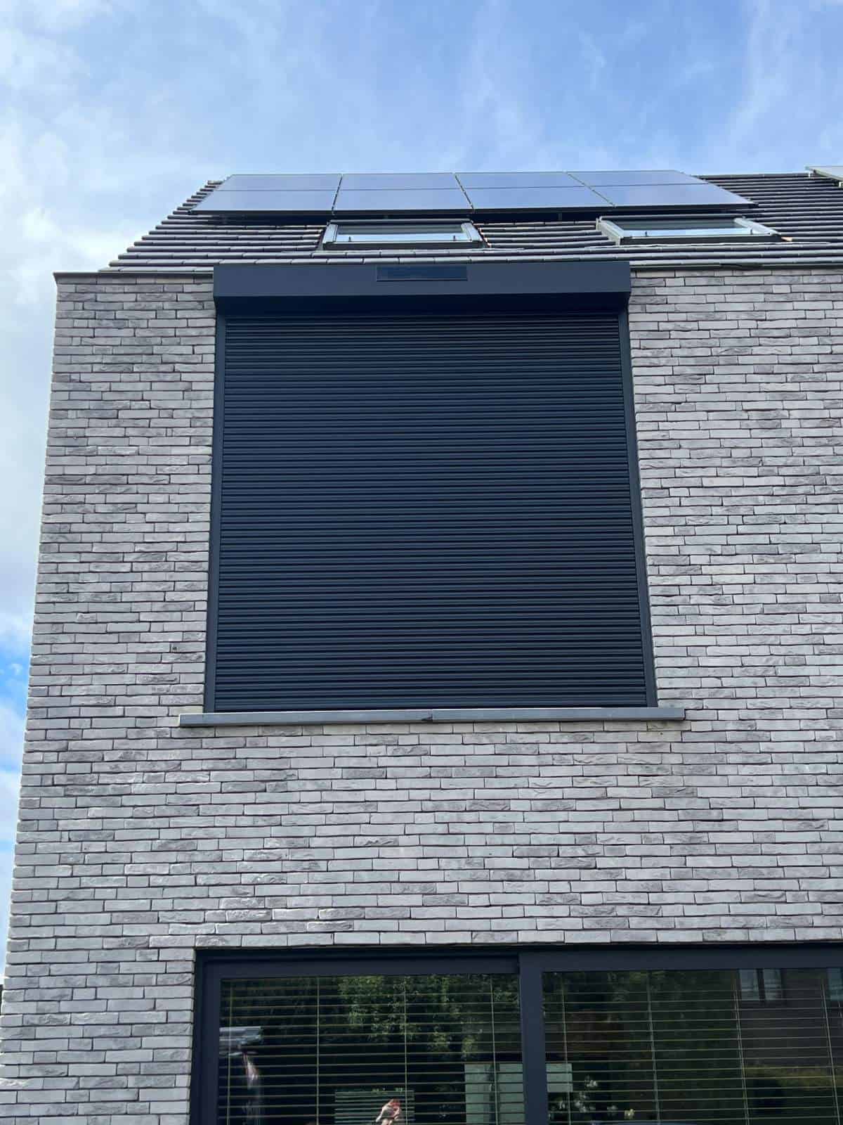 Gevel van modern lichtgrijs bakstenen huis met gesloten zwart rolluik en zonnepanelen op het dak.