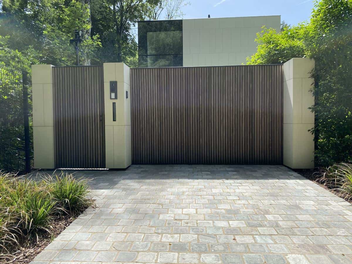 Moderne schuifpoort met verticale houten lamellen tussen hoge beige zuilen en een betegelde oprit.