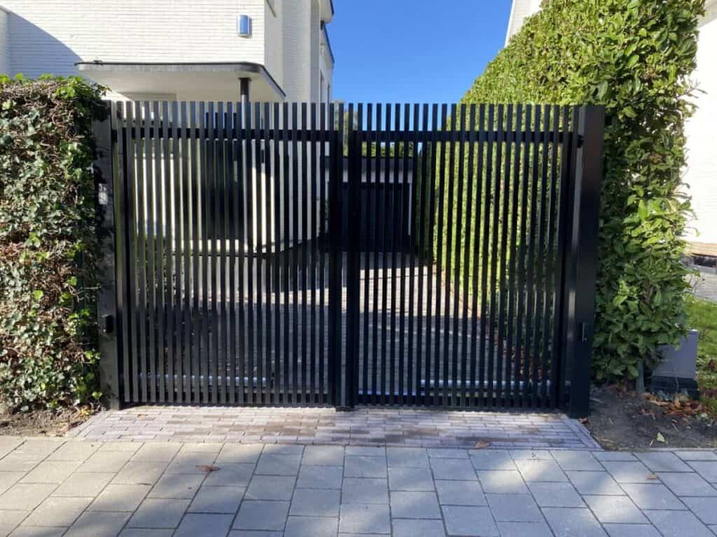 Moderne zwarte metalen draaipoort met vertical spijlen, omgeven door groene hagen naast een woning.