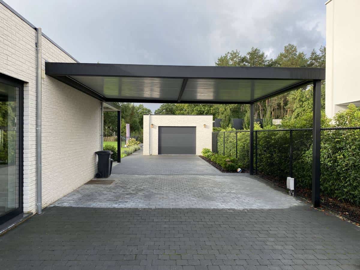 Open carport met zwart metalen dak naast wit huis, uitzicht op aparte garage en groene tuin.
