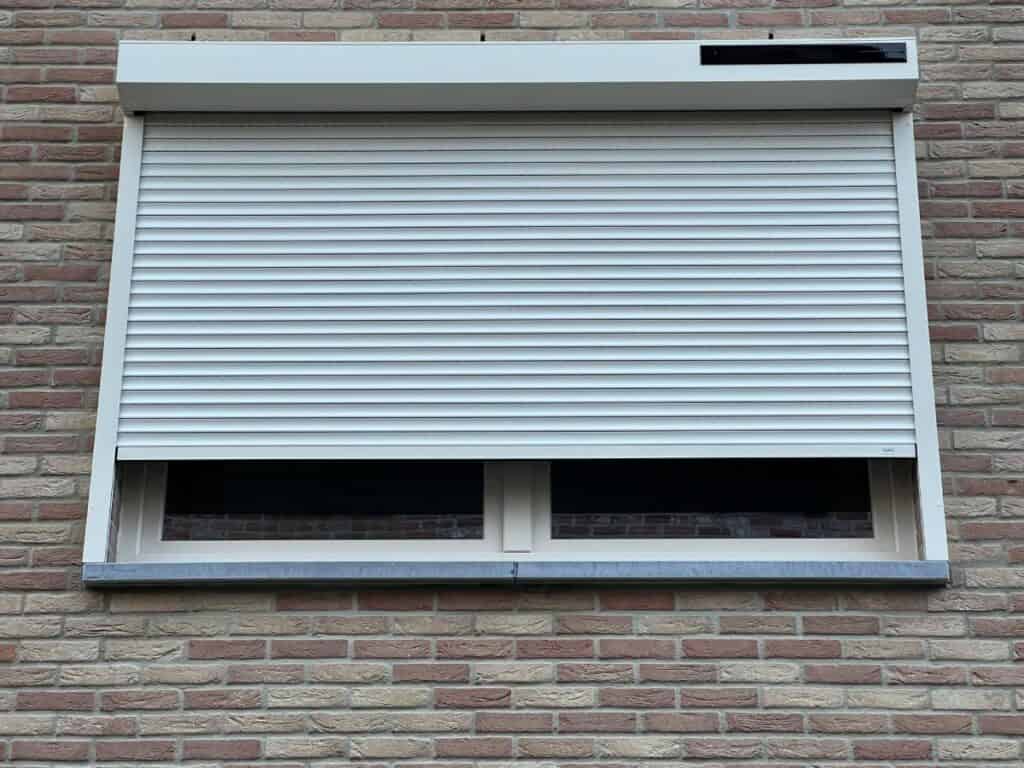 Raam in bakstenen muur met deels gesloten witte rolluik en stenen vensterbank.