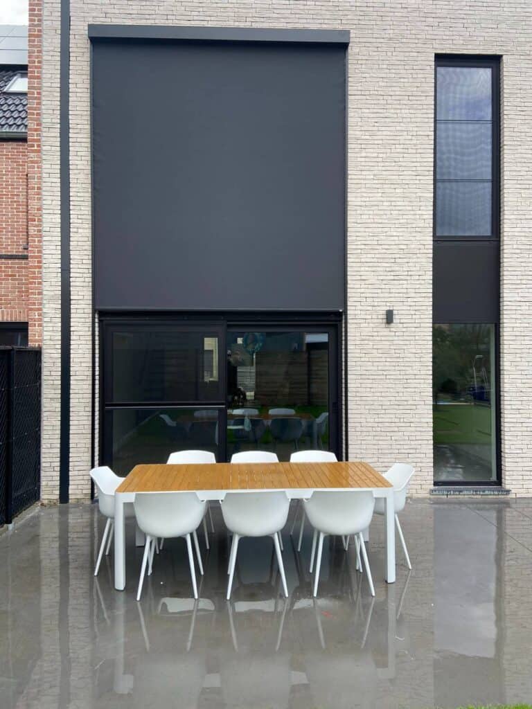 Modern terras met houten tafel en acht witte kunststof stoelen voor huis met lichte bakstenen gevel en screens.