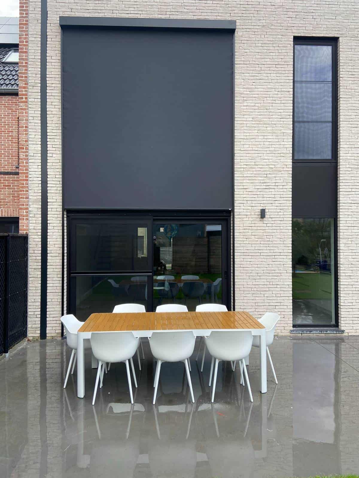 Modern terras met houten tafel en acht witte kunststof stoelen voor huis met lichte bakstenen gevel en screens.