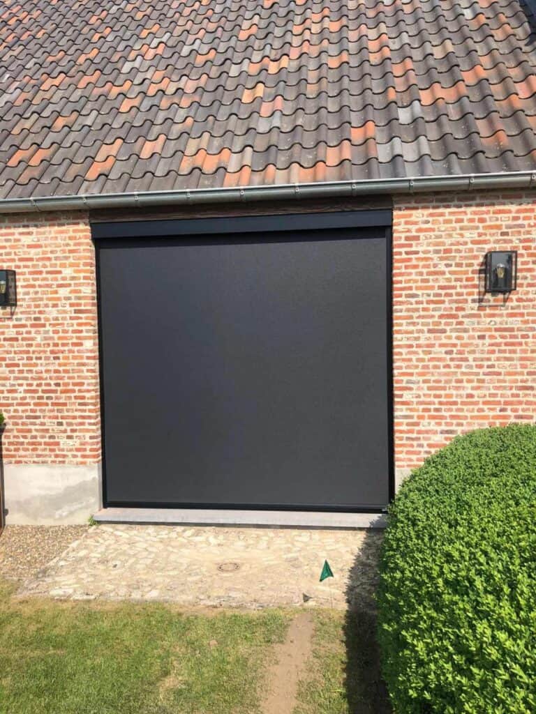 Zwarte uitschuifbare screens in bakstenen muur onder dak met dakpannen, met buitenlampen en gesnoeide struik.