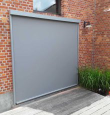 Grijs screens tegen rode bakstenen gevel met buitenlampen en planten op houten terras.